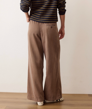 Load image into Gallery viewer, A person shown from the back wearing wide-leg brown pants, a navy and gold striped long-sleeve shirt, and white sneakers, standing against a plain off-white wall on a wooden floor.
