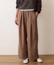 Load image into Gallery viewer, A person wearing loose, wide-leg taupe brown trousers, a navy and gold striped long-sleeve shirt, and white sneakers, standing against a plain off-white wall.