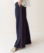 Load image into Gallery viewer, A person wearing navy wide-leg pants with a thin red and white stripe down the side, paired with a white top and white sneakers with black stripes, standing against a plain off-white background.
