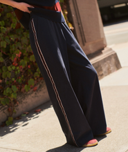 Load image into Gallery viewer, A person outdoors wearing very wide-leg navy pants with a thin light-colored stripe down the side, paired with pink shoes and a dark top. The photo is taken in natural sunlight on a sidewalk near some greenery.