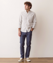 Load image into Gallery viewer, A man wearing a light beige long-sleeve button-down shirt, navy blue pants, and white sneakers, standing against a plain off-white wall.