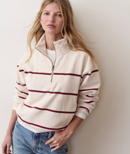 Load image into Gallery viewer, A woman wearing a cream-colored quarter-zip sweatshirt with dark red horizontal stripes, paired with blue jeans, standing against a plain light beige wall.