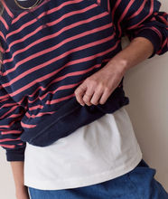 Load image into Gallery viewer, A close-up of a person wearing a navy blue and pink striped long-sleeve top over a white undershirt, paired with blue jeans. The person is holding up the hem of the striped top to show the solid navy lining.