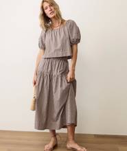 Load image into Gallery viewer, A woman modeling a matching two-piece set featuring a brown and white gingham checkered short-sleeve top with puff sleeves and a matching tiered midi skirt, standing against a plain off-white wall.