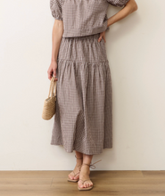 Load image into Gallery viewer, A model wearing a brown and beige gingham two-piece set consisting of a short puff-sleeve top and a tiered midi skirt, holding a small woven straw bag against a plain off-white wall.