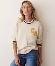 Load image into Gallery viewer, A woman wearing a cream-colored knit sweater with yellow 'CA' lettering on the chest and striped ribbed cuffs and collar, paired with blue jeans, leaning against a plain off-white wall.