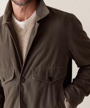 Load image into Gallery viewer, A close-up of a man wearing an olive brown utility jacket over a beige knit sweater and white t-shirt, against a plain light beige background.