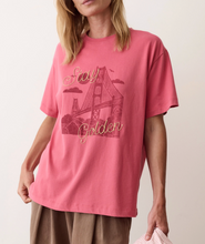 Load image into Gallery viewer, A woman wearing a pink-red short-sleeve graphic T-shirt with a faded bridge illustration and the words 'Stay Golden' in script, paired with brown pants against a plain light background.