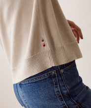 Load image into Gallery viewer, Close-up of a person wearing a cream-colored knit sweater with a small striped accent and blue jeans, photographed against a plain light background.