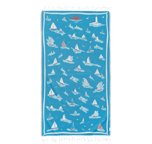 Load image into Gallery viewer, A rectangular blue beach towel with white fringe on the top and bottom edges, featuring a repeating pattern of white sailboats, small boats, and birds, with a small red sun near the top center.