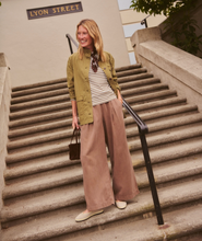 Load image into Gallery viewer, A woman stands on outdoor stone stairs wearing an olive green jacket over a striped top and wide-leg taupe pants, holding a small dark handbag. A building sign reading 'LYON STREET' is visible behind her.