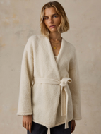 A woman wearing a cream-colored, textured knit wrap cardigan with a matching tie belt, styled with dark pants and gold necklaces against a textured beige wall.