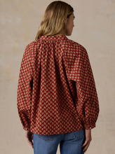 Load image into Gallery viewer, A woman viewed from the back wearing a red long-sleeve blouse with a beige paisley pattern and blue jeans, standing against a textured beige wall.
