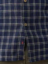 Load image into Gallery viewer, Close-up of a blue and white plaid button-down shirt, showing the fabric texture, buttons, and a small embroidered logo patch near the hem.
