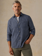 Load image into Gallery viewer, A man wearing a navy blue and white plaid button-down shirt with olive green pants, standing against a textured beige background.

