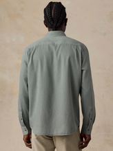 Load image into Gallery viewer, A man viewed from the back wearing a muted sage green long-sleeve button-up shirt and beige trousers, standing against a textured beige wall.
