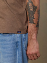 Load image into Gallery viewer, Close-up of a person wearing a brown short-sleeve t-shirt and light blue jeans, showing the side pocket seam and a small dark brand tab near the hem.
