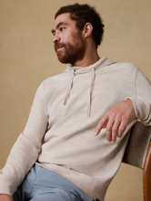 Load image into Gallery viewer, Man wearing a beige hoodie sitting on a chair against a beige background
