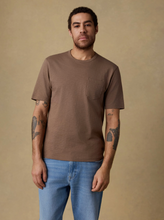 Load image into Gallery viewer, A man wearing a brown short-sleeve crewneck pocket t-shirt and blue jeans, standing against a solid beige background.

