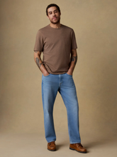 Load image into Gallery viewer, A man modeling a brown short-sleeve crewneck t-shirt and light blue jeans, standing against a solid beige studio backdrop.

