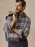Man in a tan and blue plaid button down shirt and dark pants, with a denim jacket draped over his shoulder, standing in front of a beige wall 