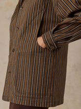 Load image into Gallery viewer, Close-up of a person wearing a brown, tan, and dark striped button-front jacket with a pocket and rolled cuff, against a textured beige background.
