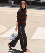Load image into Gallery viewer, A woman walking outdoors on a street crosswalk wearing a dark navy and burgundy striped knit sweater, dark wide-leg pants with a thin light stripe down the side, and red sneakers, while carrying a light blue handbag.
