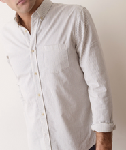 Load image into Gallery viewer, A close-up of a man wearing a long-sleeve button-down shirt with thin vertical stripes in off-white and beige.
