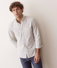 Load image into Gallery viewer, A man wearing a light beige and white micro-striped long-sleeve button-down shirt with dark blue jeans, leaning against a plain off-white wall.
