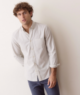 A man wearing a light beige and white micro-striped long-sleeve button-down shirt with dark blue jeans, leaning against a plain off-white wall.