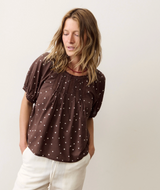 A woman wearing a brown short-sleeve blouse with white polka dots and white pants, standing against a plain light beige background.