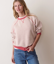 Load image into Gallery viewer, A woman wearing a light pink crewneck sweatshirt with contrasting dusty rose ribbed collar, cuffs, and hem, paired with blue jeans, standing against a plain off-white wall.
