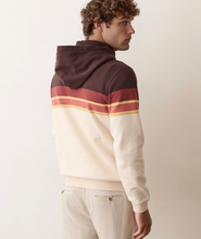 Load image into Gallery viewer, A man wearing a color-blocked hoodie with a dark brown hood and upper back, a cream body, and horizontal stripes in terracotta red and yellow across the shoulders and upper arms, paired with cream pants against a plain beige background.
