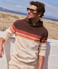 Load image into Gallery viewer, A man wearing a color-blocked hoodie and matching sweatpants stands outdoors against a white wall with a beach and ocean in the background. The hoodie has a dark brown upper section, a cream lower body, and horizontal stripes in terracotta and yellow across the chest and sleeves.
