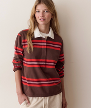 Load image into Gallery viewer, A woman wearing a brown long-sleeve rugby-style shirt with red and white horizontal stripes, paired with light beige pants, standing against a plain beige wall.
