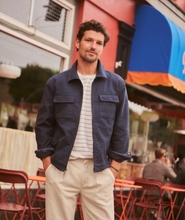 Load image into Gallery viewer, Man in a navy jacket, striped t-shirt and khaki pants outside in front of a cafe with red chairs
