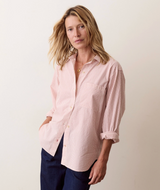 A woman wearing a light pink and white vertically striped button-down shirt with rolled-up sleeves, paired with dark navy pants, standing against a plain off-white background.