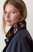 Load image into Gallery viewer, Close-up portrait of a woman wearing a dark navy jacket with a contrasting plaid collar in beige, brown, and burgundy tones, against a plain off-white background.
