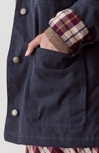 Load image into Gallery viewer, Close-up of a dark navy jacket with a large front patch pocket, metal snap buttons, and a hand tucked into the pocket. A brown ribbed knit sleeve with a burgundy and tan plaid cuff is visible.
