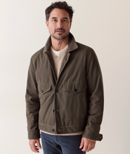 Load image into Gallery viewer, A man wearing an olive green utility jacket over a beige knit sweater and blue jeans, standing against a plain off-white background.
