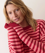 Load image into Gallery viewer, A woman wearing a red and pink striped knit sweater, posing with her hand on her chin against a plain light background.
