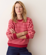 A woman with blonde hair wearing a red and pink striped knit sweater and dark blue jeans, standing with arms crossed against a plain off-white wall.