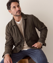Load image into Gallery viewer, A man wearing an olive green utility jacket over a beige sweater and blue jeans, sitting on a wooden stool against a plain beige wall.
