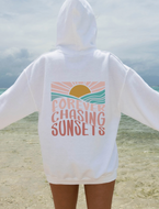A person standing on a beach with their back to the camera, wearing a white hoodie with a retro sunset graphic and the text 'FOREVER CHASING SUNSETS' printed on the back.