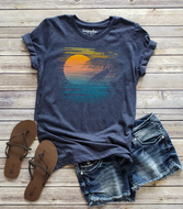 A navy blue graphic t-shirt with a sunset beach print, styled in a flat lay with denim shorts and brown sandals on a light wood plank background. 