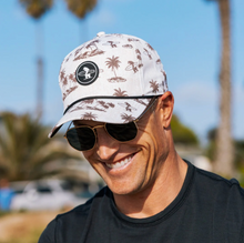 Load image into Gallery viewer, A man wearing a white baseball cap with a brown palm tree and surfboard pattern, black sunglasses, and a black t-shirt, photographed outdoors with a sunny blue sky and palm trees in the background.
