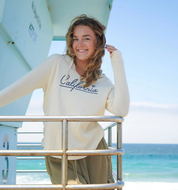 A woman smiling and leaning on a metal railing on a boat, wearing a cream-colored hoodie with 'California' written across the chest in navy script, with the ocean in the background.